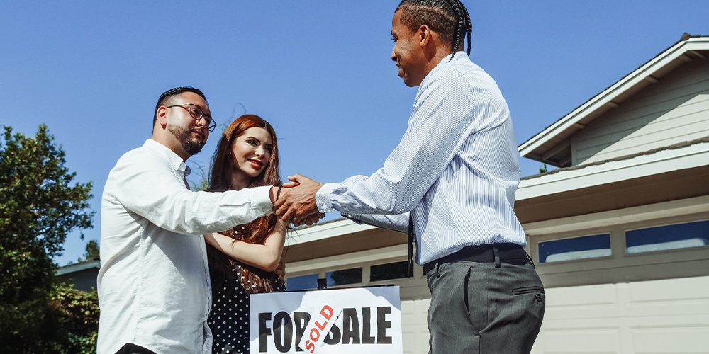 Real estate agent shaking hands with clients over a For Sale sign marked sold.