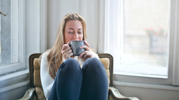 A white woman relaxes in a seat in a corner by some windows, managing stress by sipping on a cup of a nice beverage.