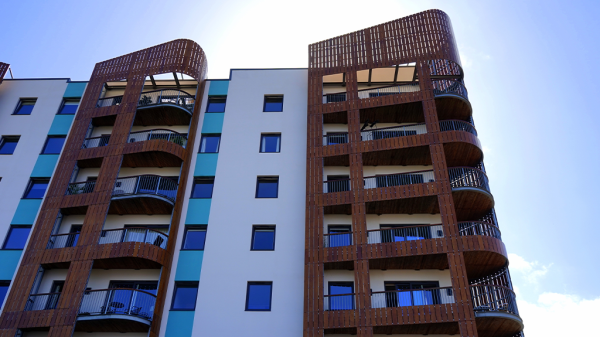 A ground view looking up at a modern set of apartment housing with the sun glowing behind it.