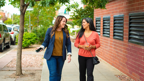 Two women walking down the street in an active conversation. One woman holds a phone in her hand while she considers giving her phone number.