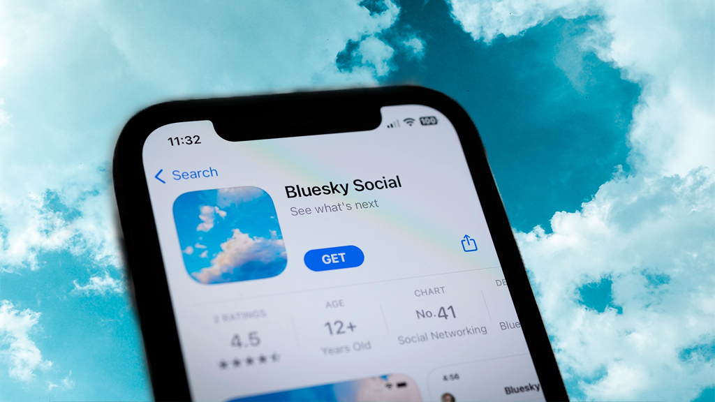 A smartphone held up to a blue sky with clouds, open to the Bluesky social media app download page.