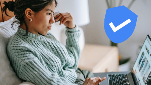 A woman sits on a couch with a laptop open, browsing with a thoughtful expression. A blue verified check mark in a blue shield shape sits on top of the scene.