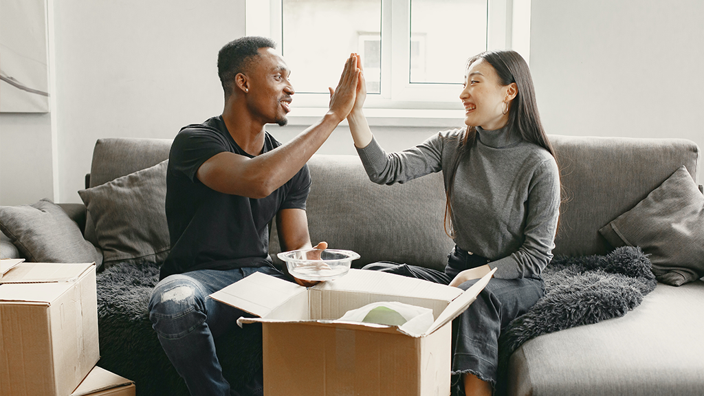 A Black man and an Asian woman high five on a couch as they unpack from their homeownership journey.