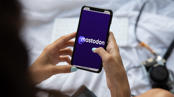 A woman holds a phone open to Mastodon app while she sits on a white bed.