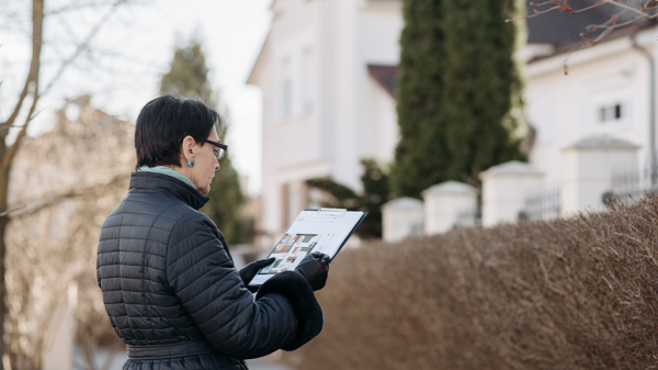 A woman stands in front of a house holding a clipboard as she reviews the documents of home sales.