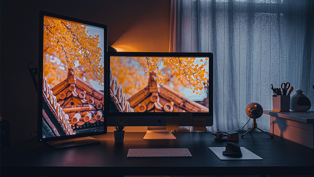 A set of two desktop monitors displays a brick house and orange leaves behind it, showing digital real estate on their computer sitting on top of a modern dark desk.