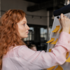 A woman with long red hair stands at a marketing board with multiple post-it notes, manually writing what AI services can assist with.