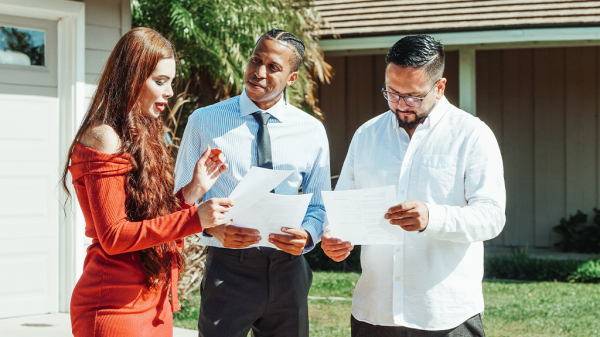 A group of three people stand outside a house with a Realtor in the middle, helping the other two to read the forms of the Nosalek case.