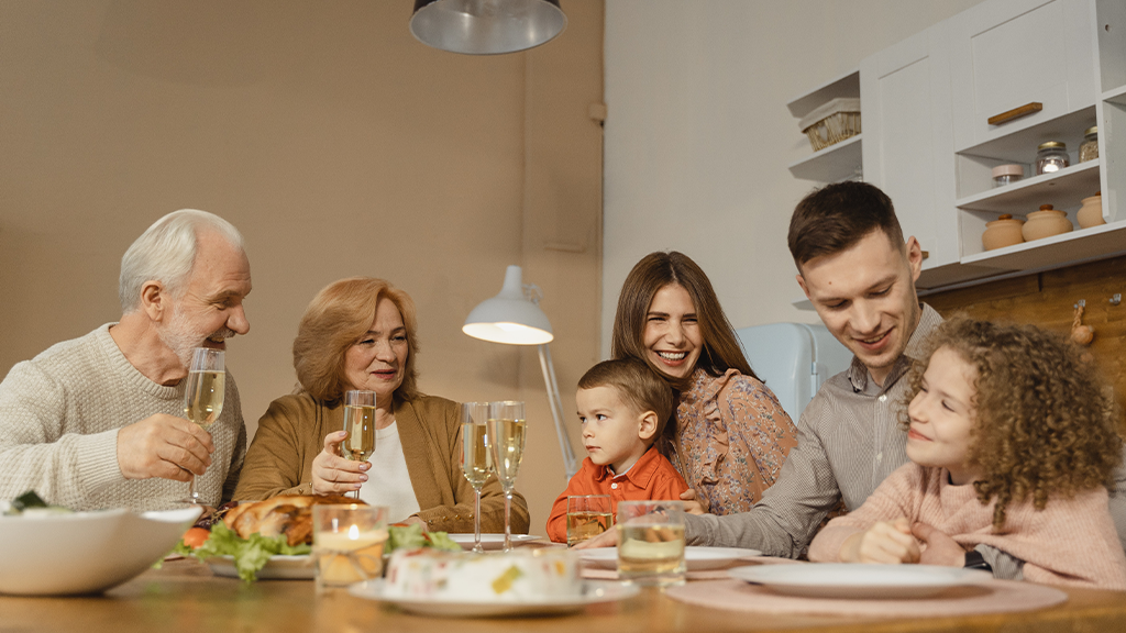 A family with grandparents, parents, and young children, all toasting and talking around a kitchen table, showing a difference in homebuying.
