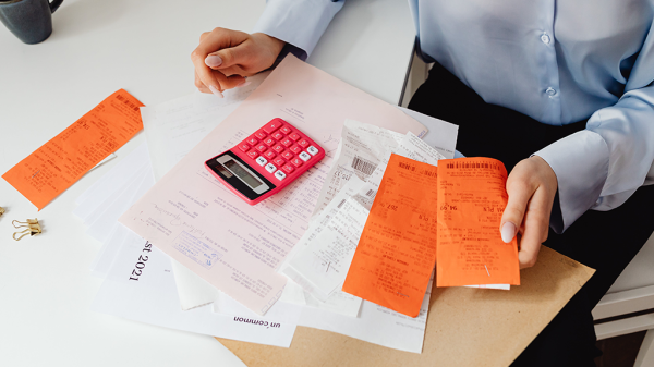 A table of reciepts, white and orange papers relating to homeownership, and a person using a pink calculator in a top down view of their table.