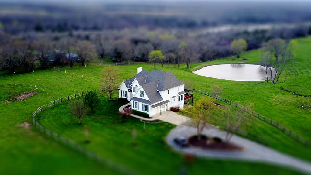 An overhead view of a large country home surrounded by green fields, with trees in the field beyond, on prime real estate.