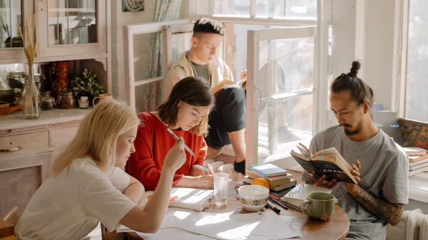 A group of young adults sitting around a kitchen table in a co-owning situation working on their own work and hobbies. One man reads at the table, another sits at the window seat in the back, one woman works on a laptop at the table, and another writes in a journal at the table.