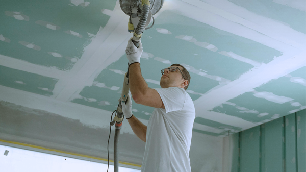 A construction worker sanding the interior ceiling of a house to avoid an OSHA violation.