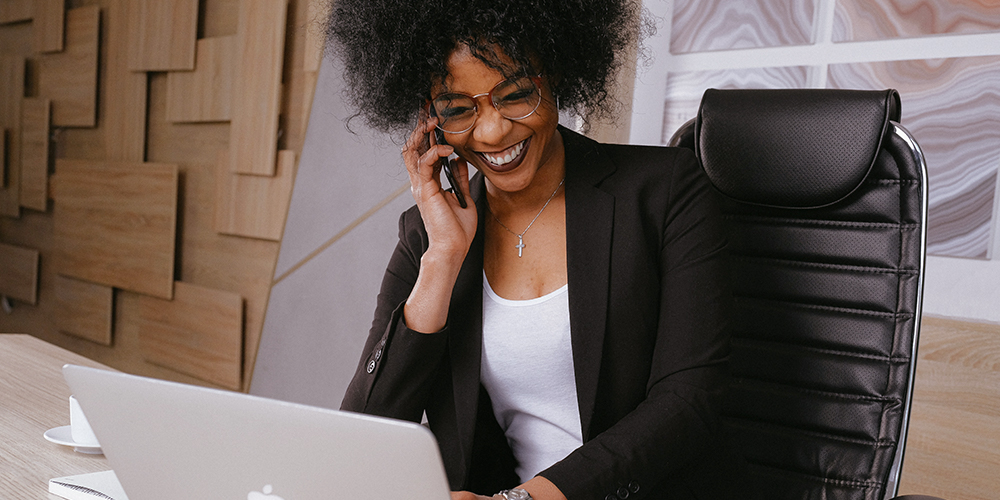 Black woman smiling in communication talking on phone and laptop in front of her.