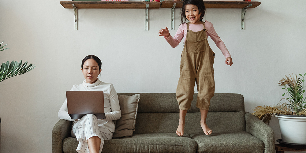Mother working remotely with a child jumping on the couch next to her working.