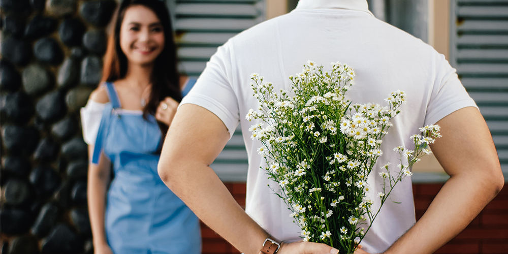 Man holding flowers behind his back while woman in background smiles. Background checks can ensure a safe modern dating experience.