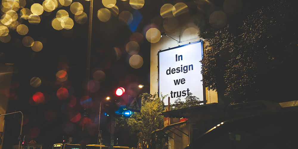 A white sign in an urban setting reading "In Design We Trust" with glowing yellow lights above.