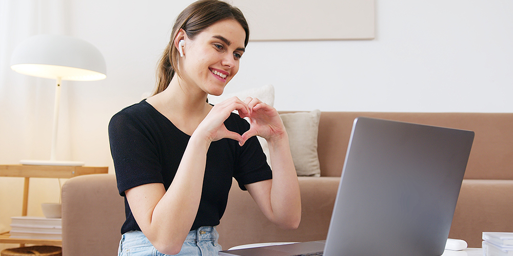 Woman forming hands into heart shape at laptop hosting live video chat, similar to Facebook's new app Hotline