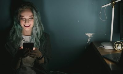 Smiling woman seated in dark room illuminated by lamp and phone light, participating in audio chat room.