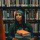 African American woman in library with two books and graduation cap.
