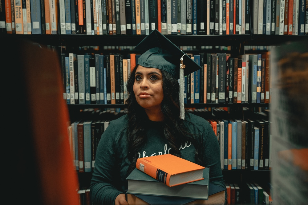 African American woman in library with two books and graduation cap.