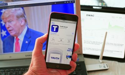 Man holding iPhone with Truth Social app download page up, as well as the stock market and Trump in the background on computer screens.