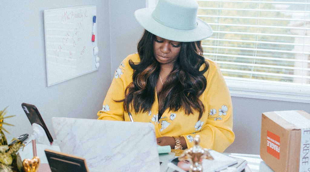Woman working at her desk with hat and yellow top representing Metaverse fashion.