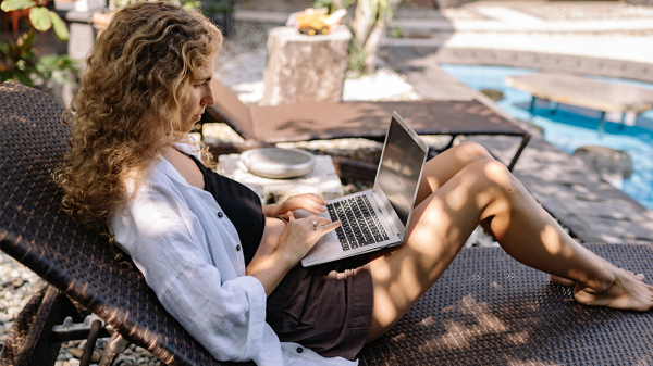A woman lounges on an outdoor chair beside a swimming pool, working on a laptop to work smarter.