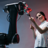 A robot AI arm reaches out toward a woman who offers a flower in return. They stand against a blue and red illuminated background.