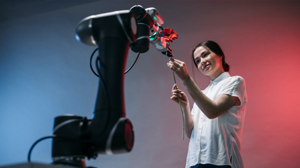 A robot AI arm reaches out toward a woman who offers a flower in return. They stand against a blue and red illuminated background.