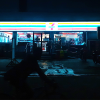 A nighttime view of a 7/11, where only the storefront is illuminated. A person rides their bike in front of the store on their way past.