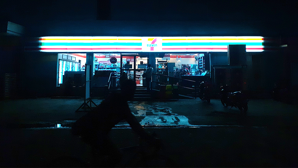 A nighttime view of a 7/11, where only the storefront is illuminated. A person rides their bike in front of the store on their way past.