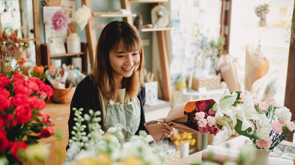 A woman surrounded by flowers in a flower shop looks down at her freelancing computer and smiles.