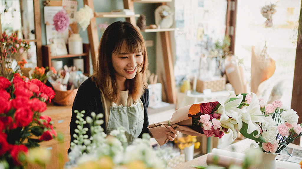 A woman surrounded by flowers in a flower shop looks down at her freelancing computer and smiles.