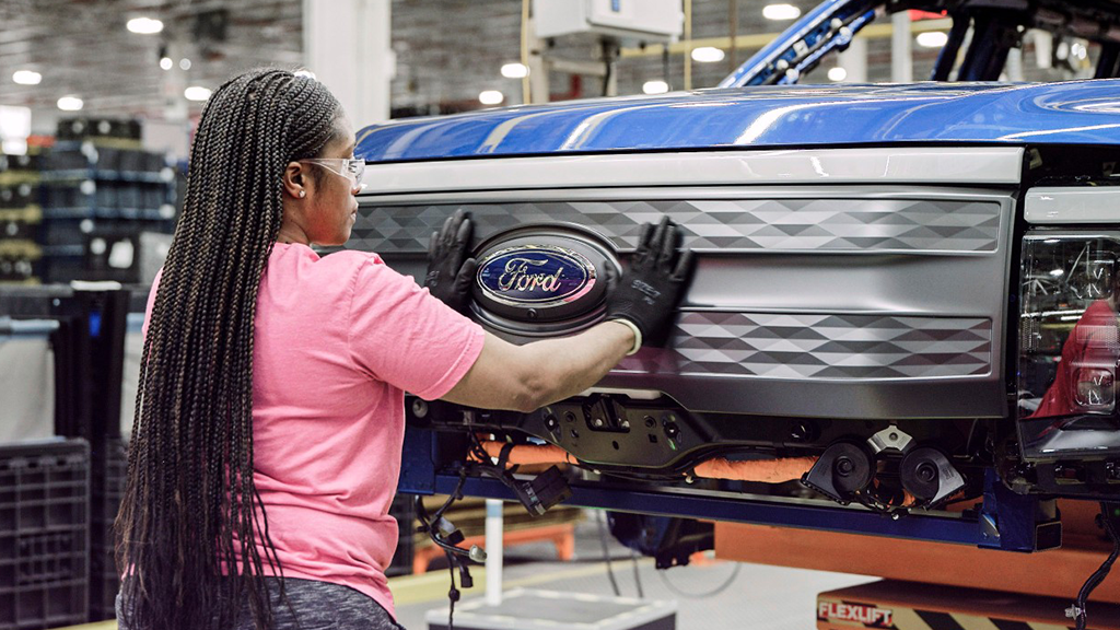 A Black woman working in a factory presses the front grill onto a Ford electric vehicle.