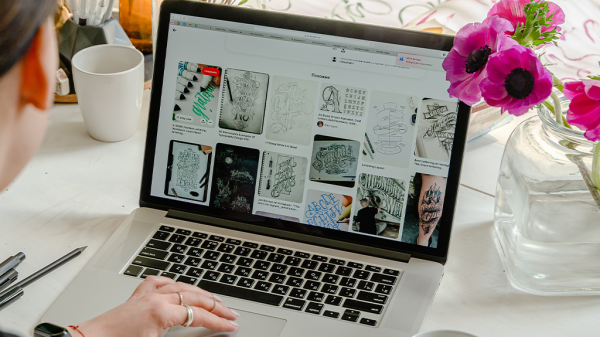 A laptop on a white desk is opened to Pinterest pins, with a person browsing them. A vase of pink flowers sits to the right of the computer.