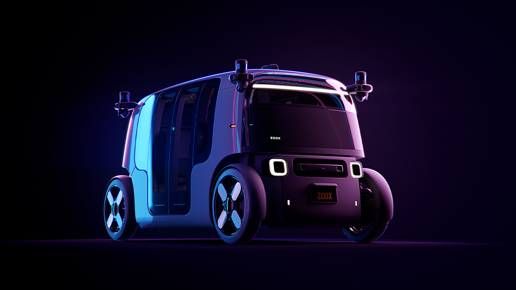 A three-quarters view of Zoox, Amazon's new robotaxi. It looks like a single tram compartment, with only passenger seats and no driver, and round wheels that can go in all directions. It is illuminated here in blue and purple light.