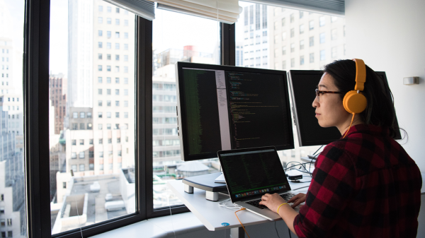 A woman in plaid and wearing yellow headphones looks out of the window as multiple computer monitors with coding on the screen await input from chatGPT and others.