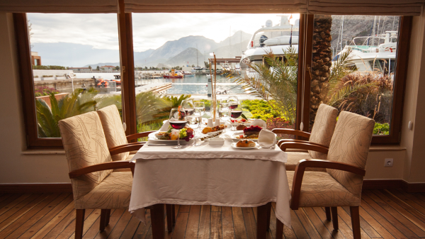 A luxury dining room with food on the table and the view of a ship outside of the window beyond it.