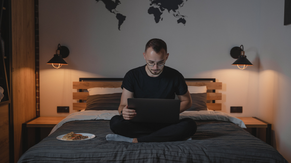 A hybrid remote worker sits on a bed in a bedroom working on a laptop, with a plate of food sitting nearby.