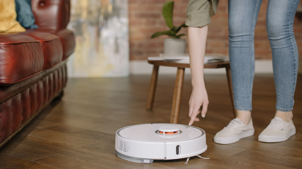 A woman inside a modern home with wooden floors and a leather couch bends over a robot vacuum to press a button on top of it. Amazon Alexa may work with one of these soon.