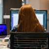 A person with long red hair sits in an office with three computers open in front of them with different coding programs and ChatGPT open.