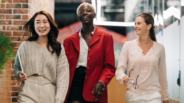 A trio of confident qualified women walk together in an office environment, with one Asian woman, a Black woman, and a white woman and talking and laughing together.