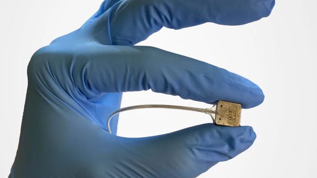 A hand wearing surgical gloves holds up a clean brain implant device, a small square with wires coming out of it.