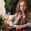 A woman sitting at a table looking up search advertising on her smart phone with a foamy coffee next to her in a cafe with various greenery.