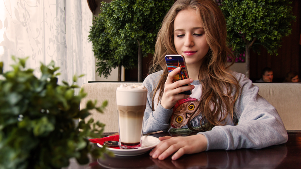 A woman sitting at a table looking up search advertising on her smart phone with a foamy coffee next to her in a cafe with various greenery.