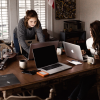 Two women sit in a cluttered, lived-in dining room working on their laptops, combating imposter syndrome in their own way.
