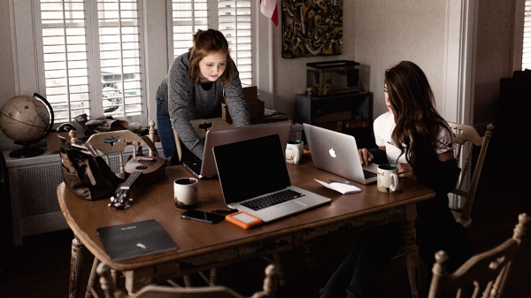 Two women sit in a cluttered, lived-in dining room working on their laptops, combating imposter syndrome in their own way.