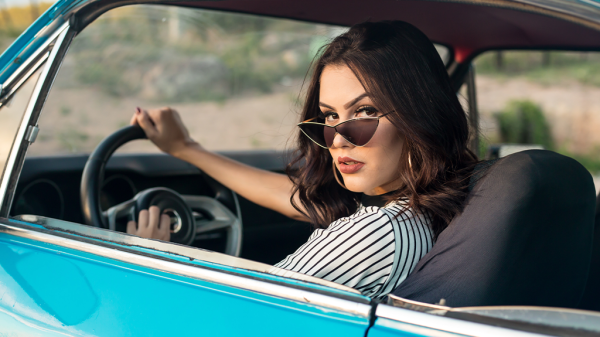 A woman wearing stylish sunglasses, sitting in the driver's seat looking back toward the camera, with her hand over the car horn.