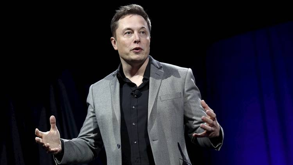 Elon Musk, a tall white man with black hair, stands on a stage gesturing as he speaks.